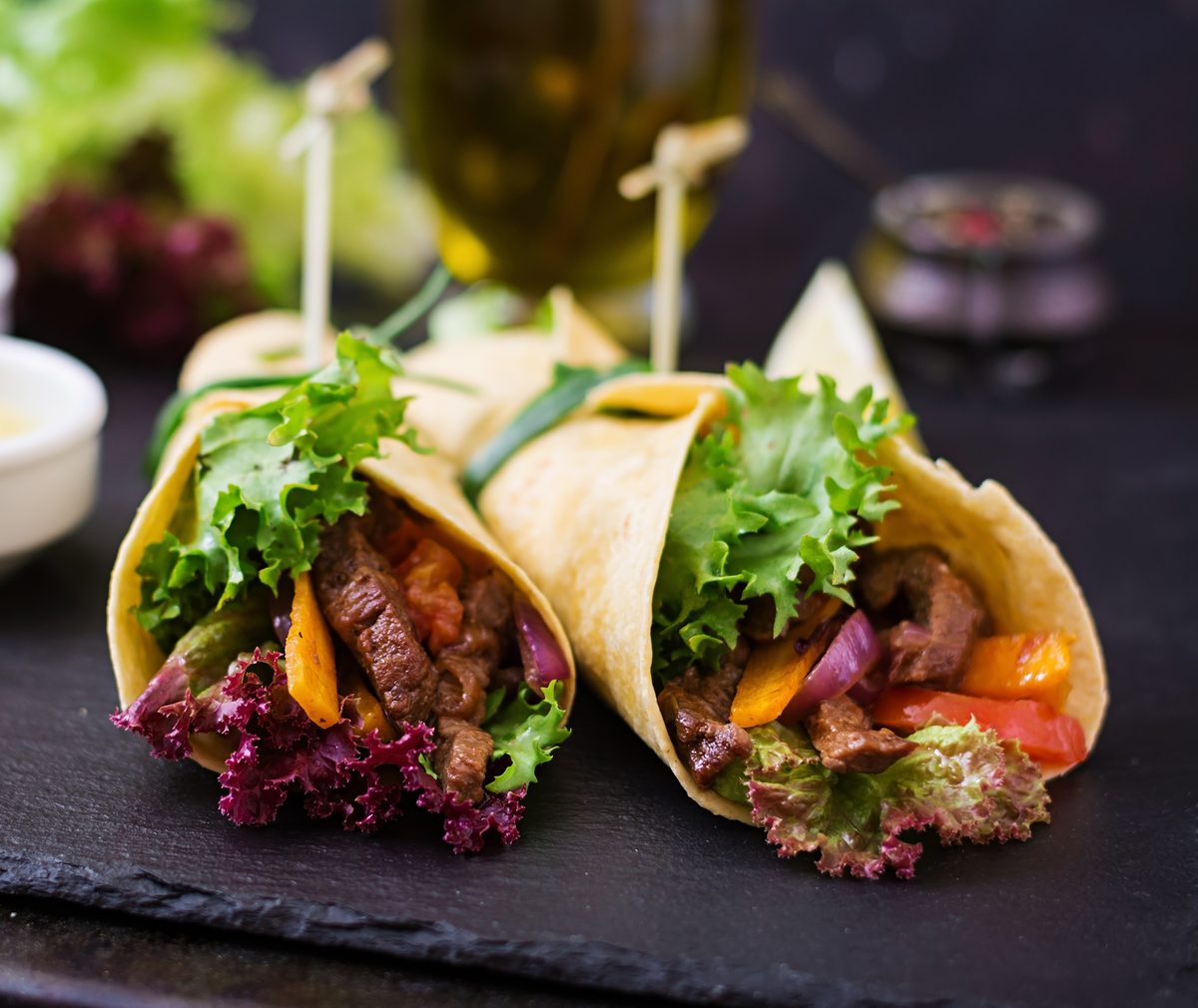 Steak wraps with peppers and lettuce on a dark slate board.